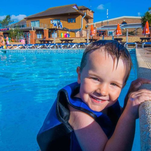A happy child wearing a life jacket in an outdoor pool with picnic benches and a flume in the background