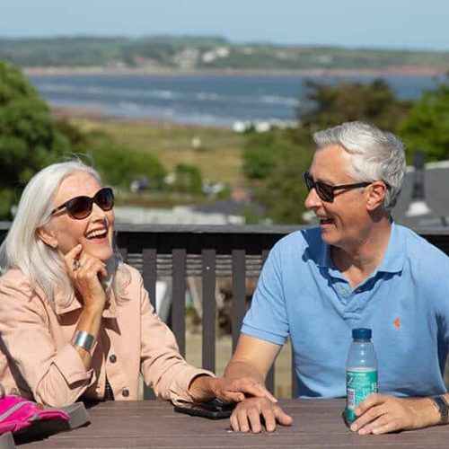 An elderly couple laughing whilst sat on a table in the sun